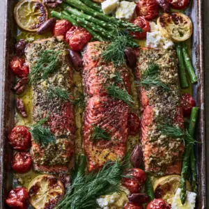 Close-up of three thick Atlantic salmon fillets with an herb crust on a sheet pan. (Mediterranean Baked Salmon Recipe)
